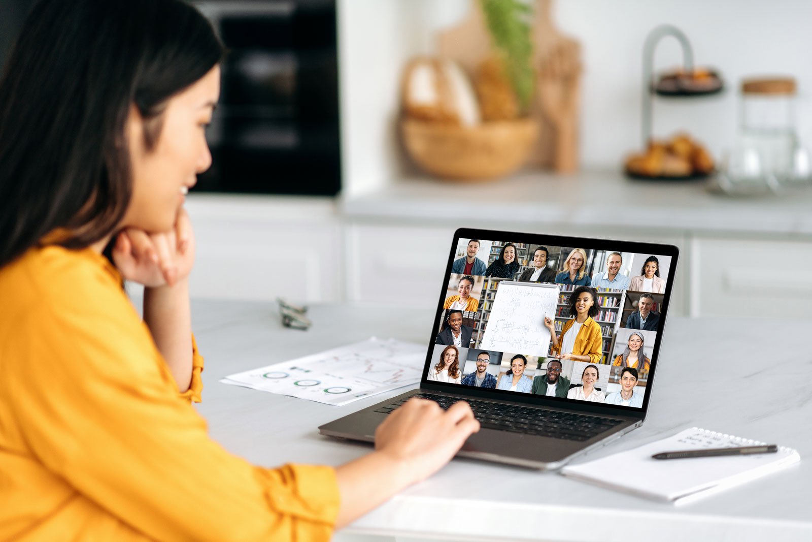 Remote lesson. Positive smart Asian female student, sitting at the table, listening to an online lecture, on the screen of a laptop, a teacher telling information, and a group of multiracial people